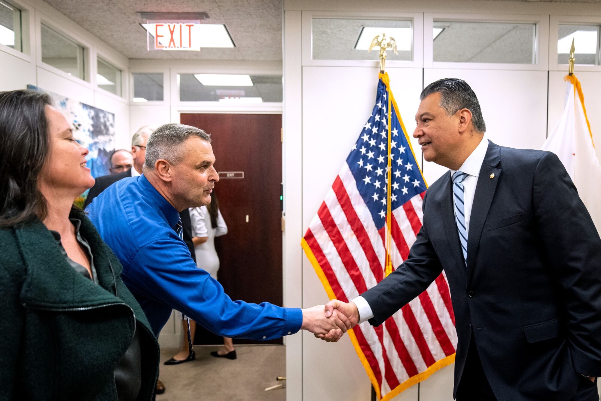 Two men shake hands while an American flag hangs on a stand behind them.