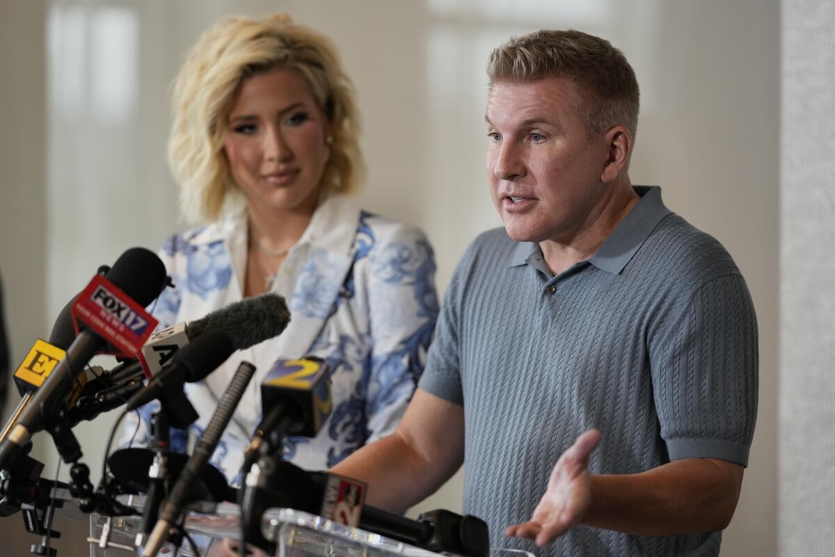 A young blond woman looks on as her father stands at a lectern and speaks into a bank of news microphones