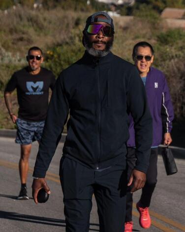 A man walks in black running clothes, with two other men walking behind him