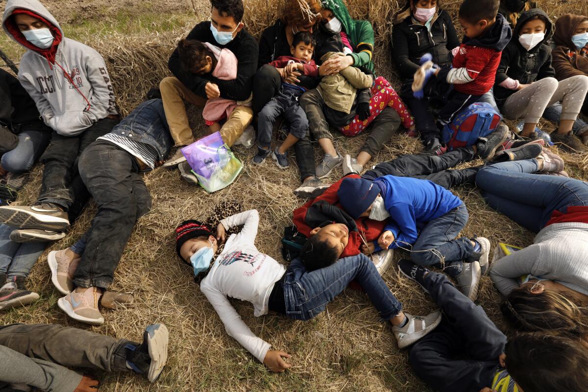 A group of migrant adults and children rest on dry grass