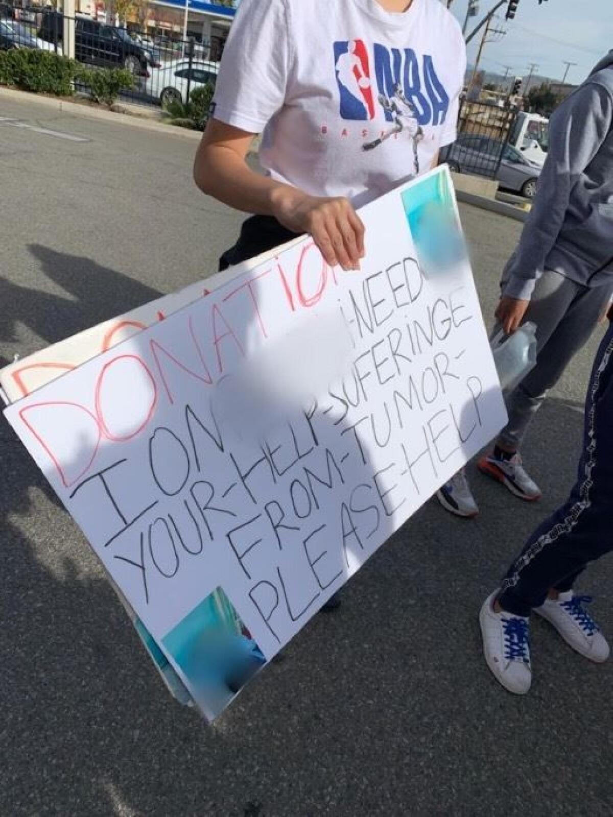 A teenager holding a handwritten sign asking for donations for someone suffering from a tumor