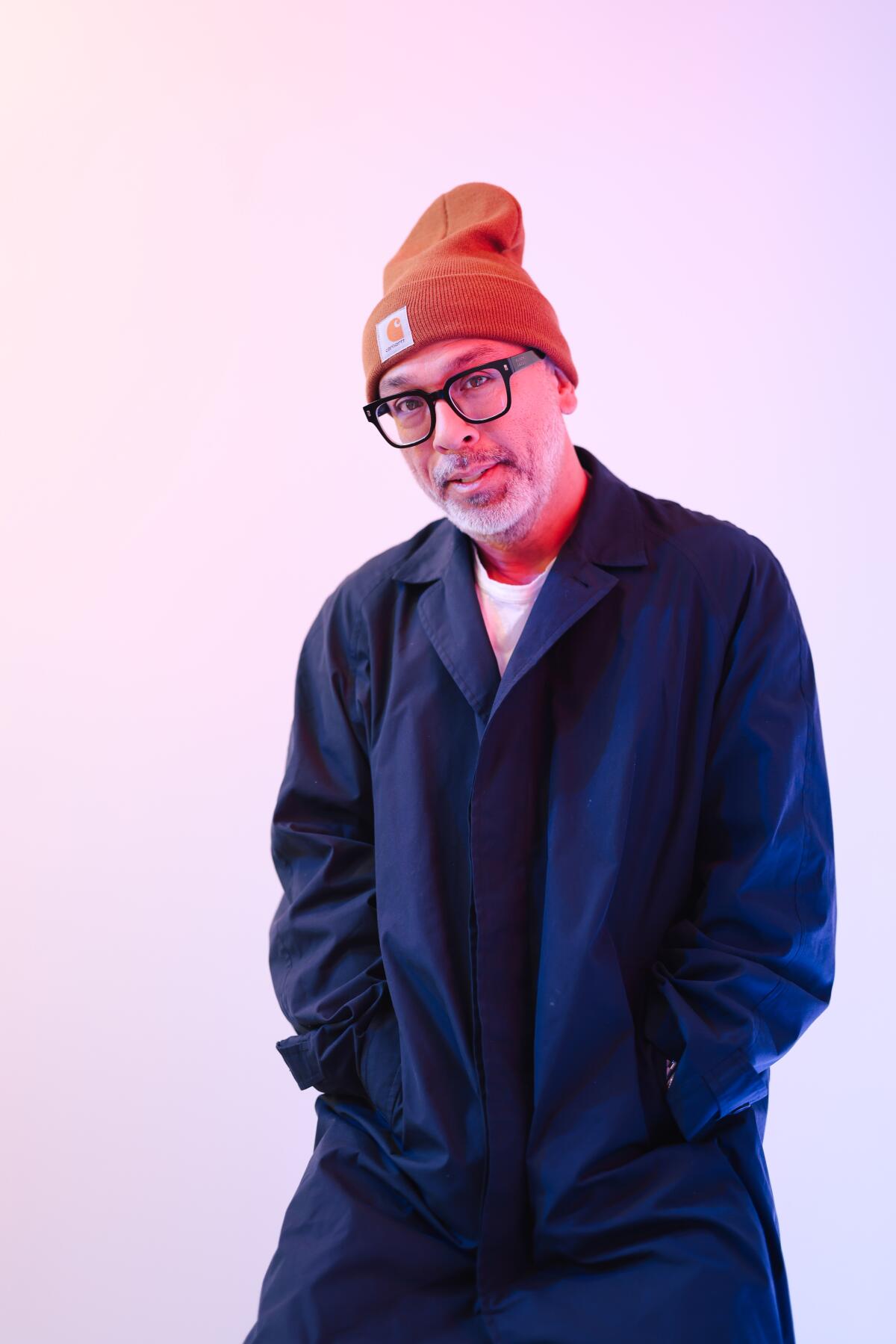 A man in a beanie, glasses and long coat looks into the camera.