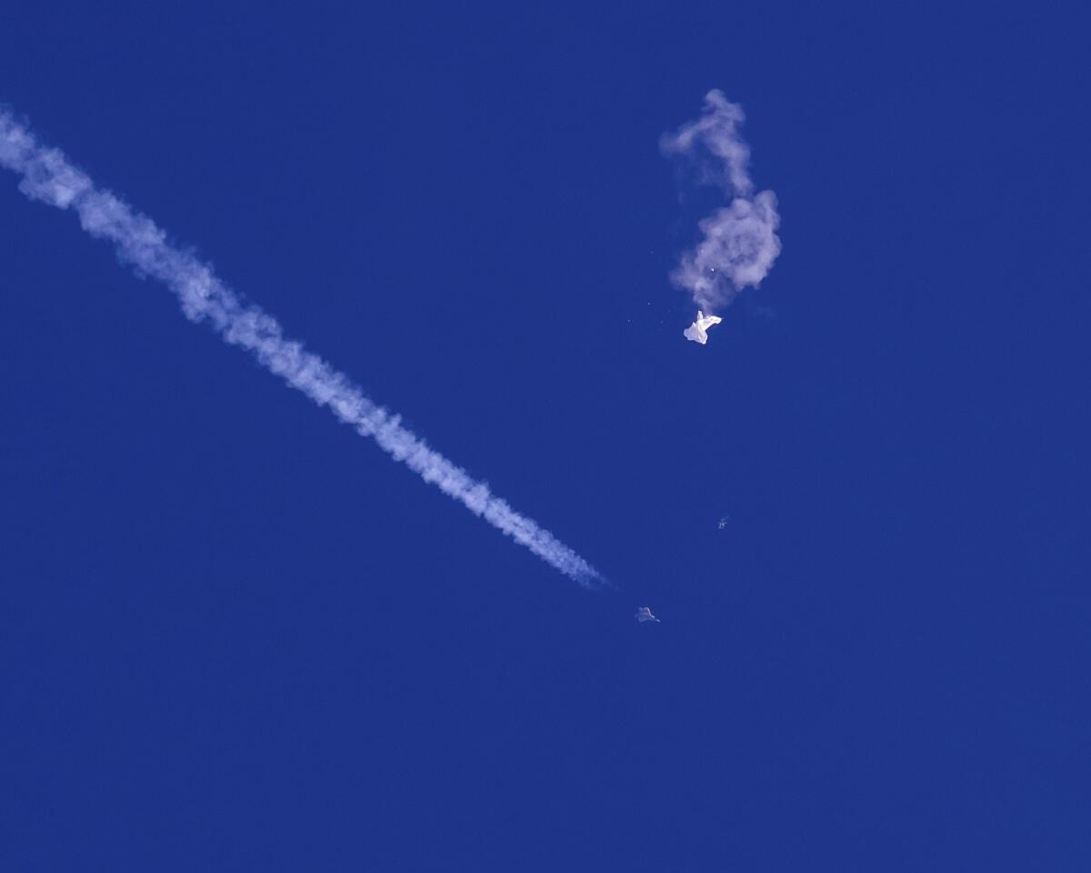 The remnants of a large balloon drift above the Atlantic Ocean with a fighter jet and its contrail below it.