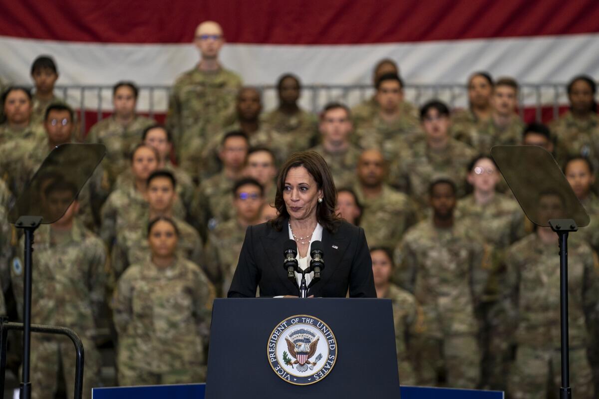 A woman stands at a lectern with people in camouflage behind her.