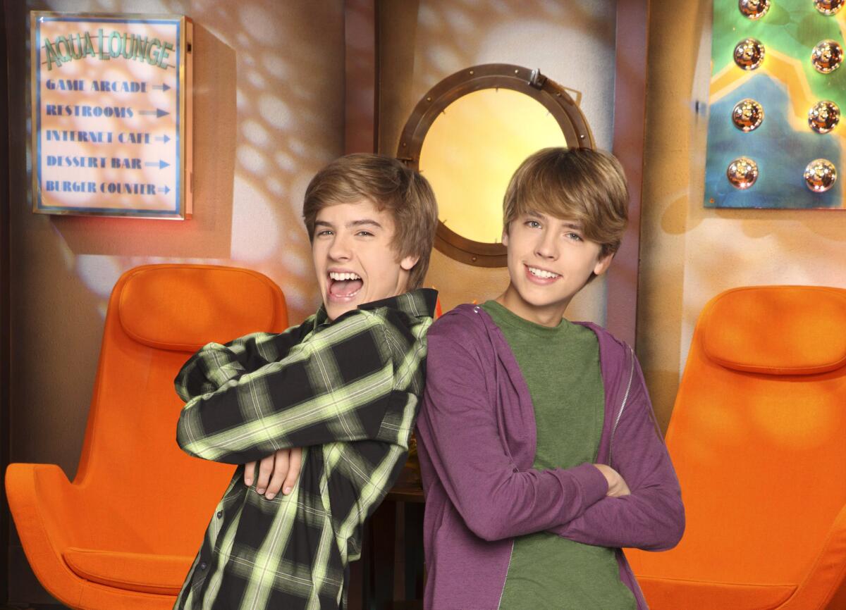 Dylan Sprouse and Cole Sprouse stand back-to-back in front of orange chairs