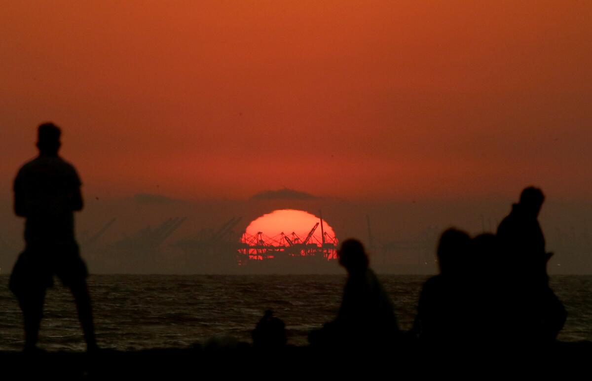The sun sets behind the Port of Long Beach with people in the foreground on the beach.