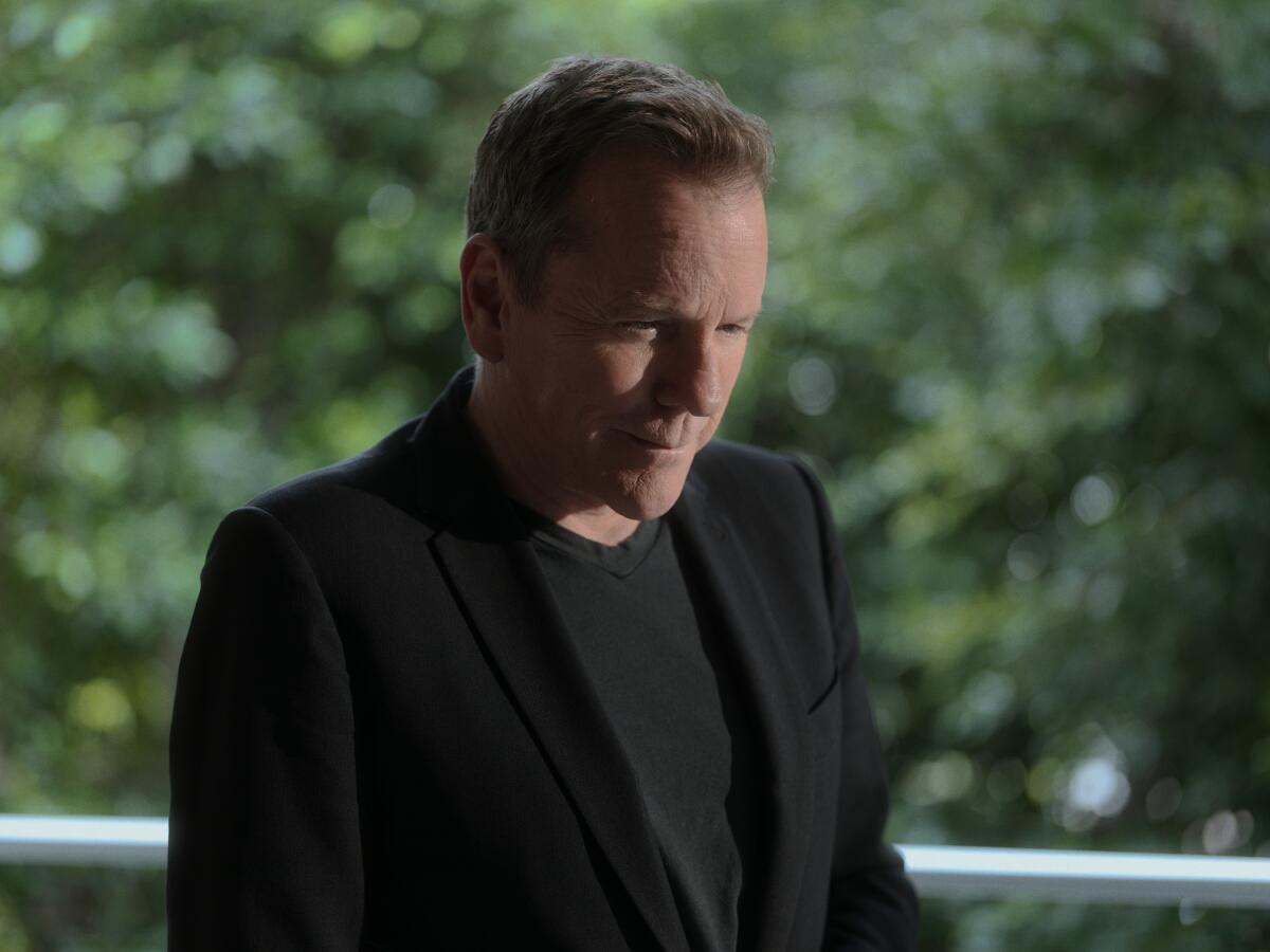 Kiefer Sutherland, in a dark shirt and blazer, stands outside looking down for a portrait.