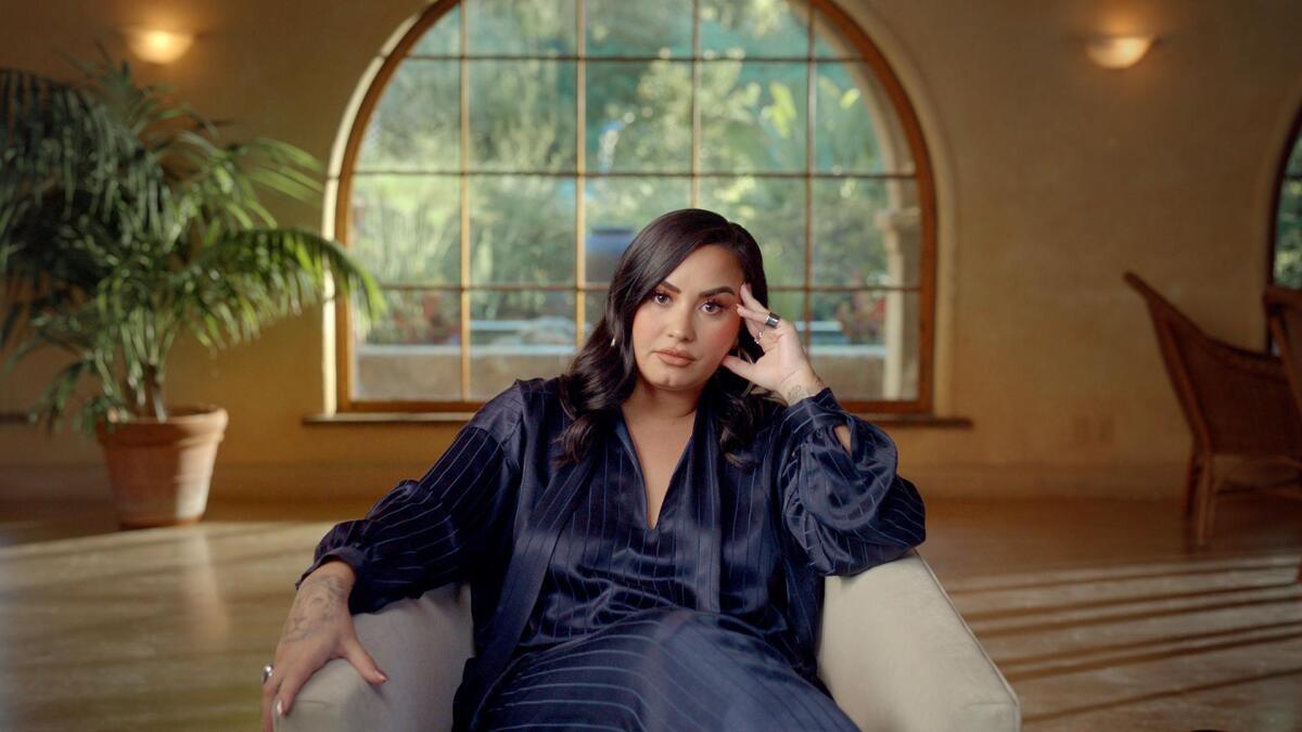 Demi Lovato wearing a silk outfit, sitting in a white armchair 