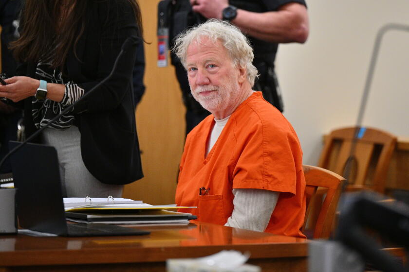 Director and actor Timothy Busfield in an orange jumpsuit sits behind a desk in a courthouse.