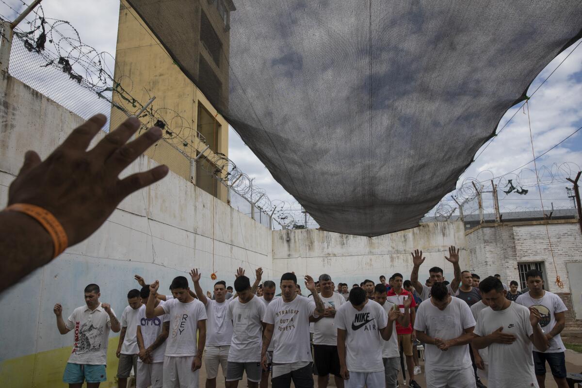 Inmates  standing in a prison yard pray, some holding up their hands.
