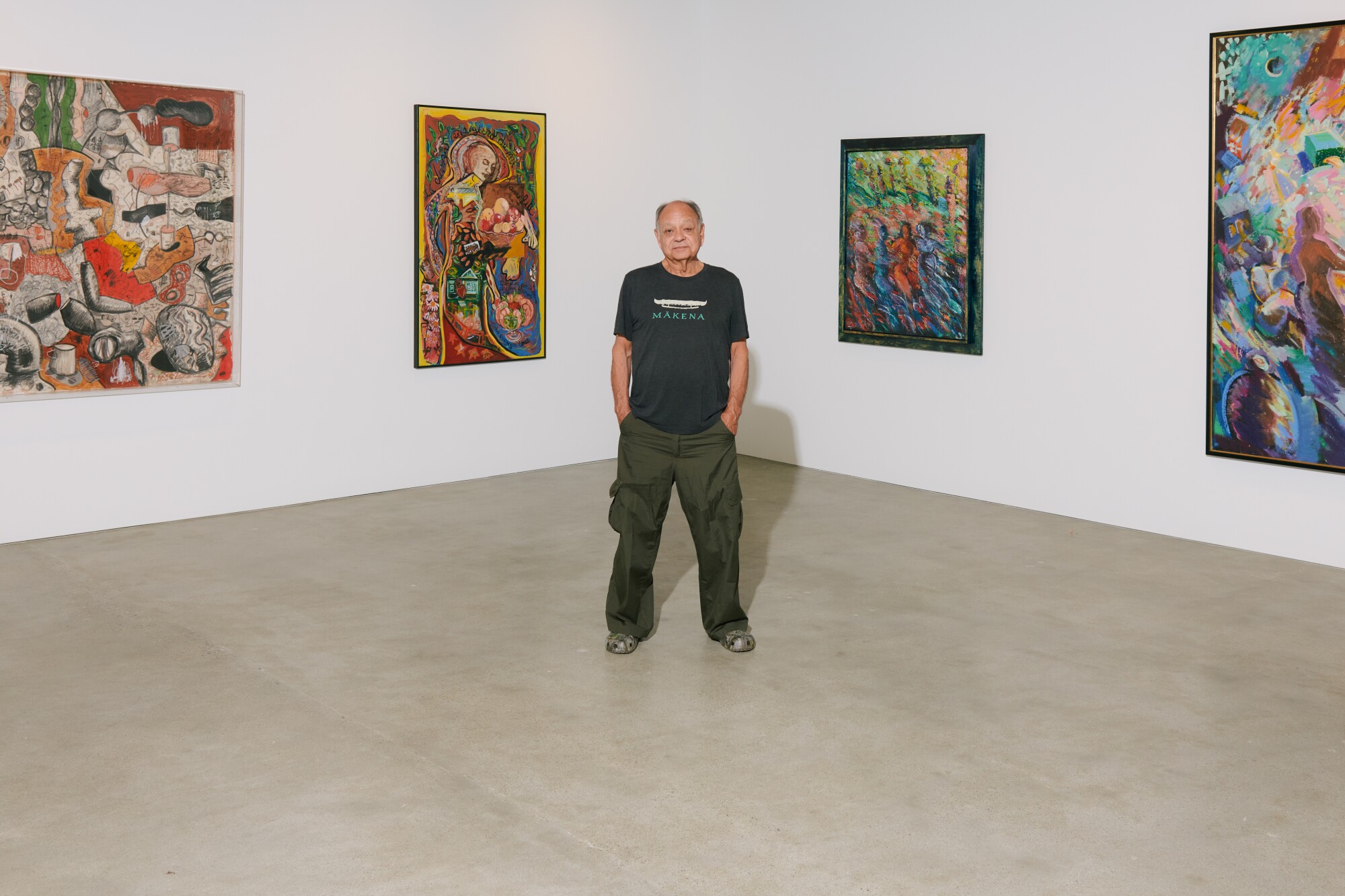 A man stands with his hands in his pockets in front of colorful paintings in a gallery with white walls.