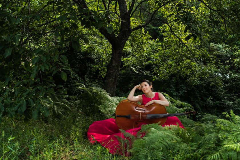 Andrea Casarrubios in a red dress sits in a forest with her cello.