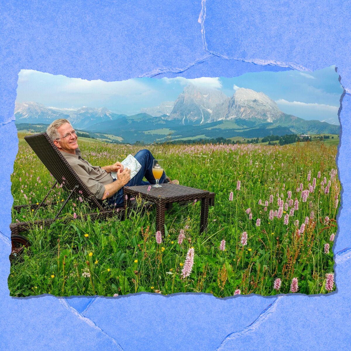 A man leans back in a chair, a drink at his side, amid a field of flowers with a view of majestic mountains.