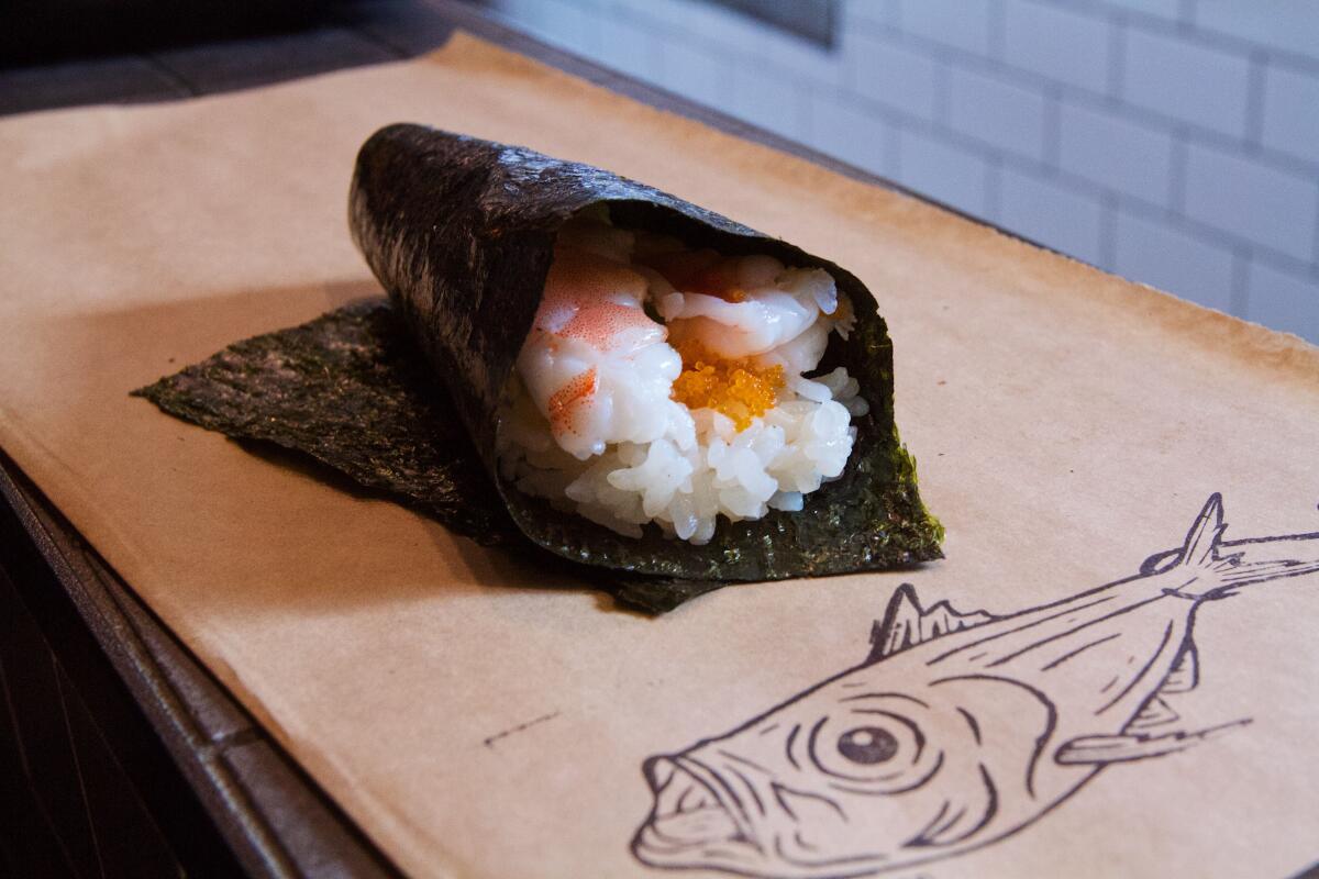 A photo of a prawn and tobiko hand roll on the counter atop brown paper featuring a line sketch of a fish.