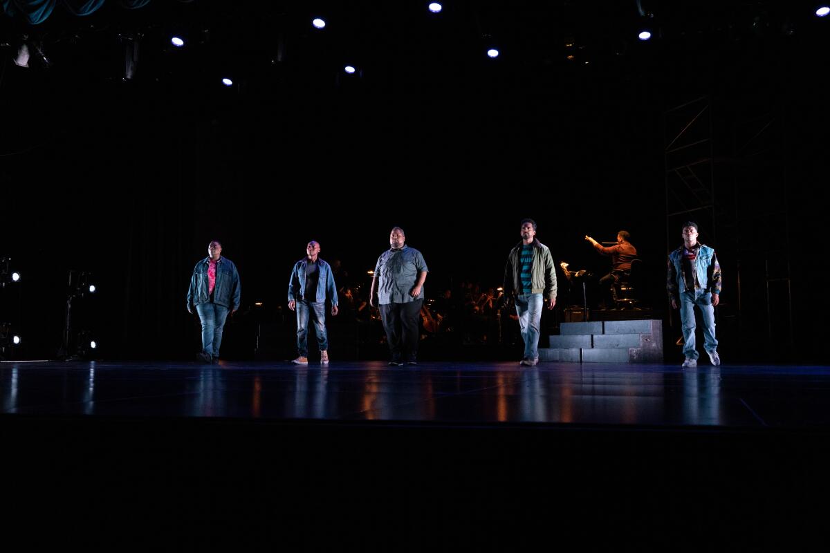Five performers stand on a dimly lighted stage that features only a low riser and the orchestra behind them.