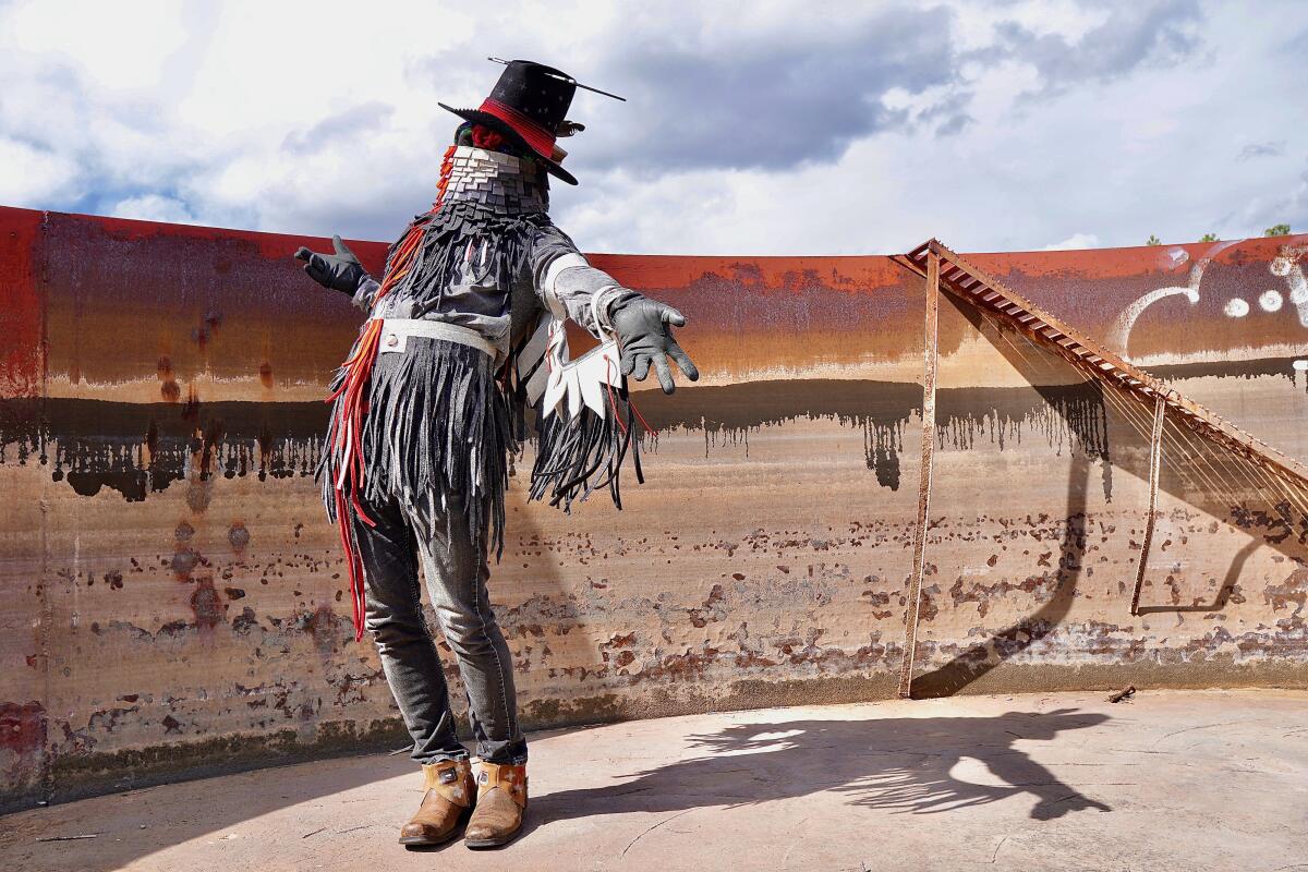 A masked man in black and white fringed wear opens his arms to the sun.