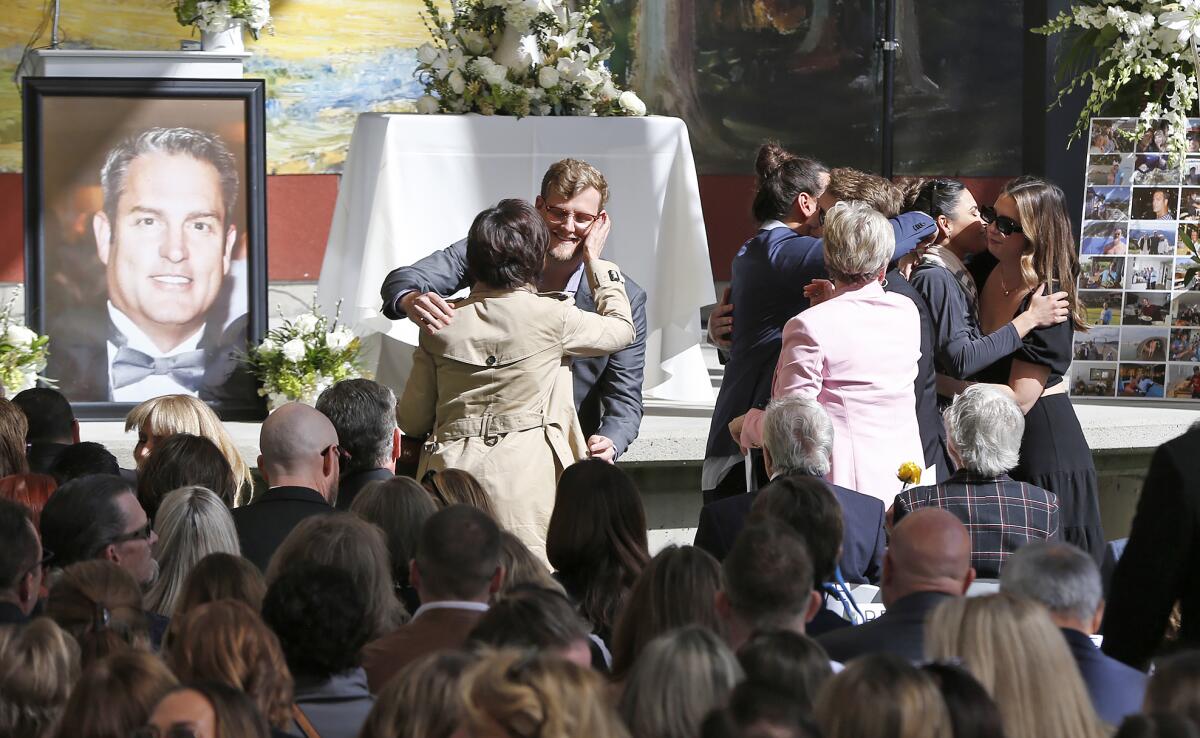 A  portrait of Michael Mammon stands next to the stage as mourners embrace during outdoor ceremony