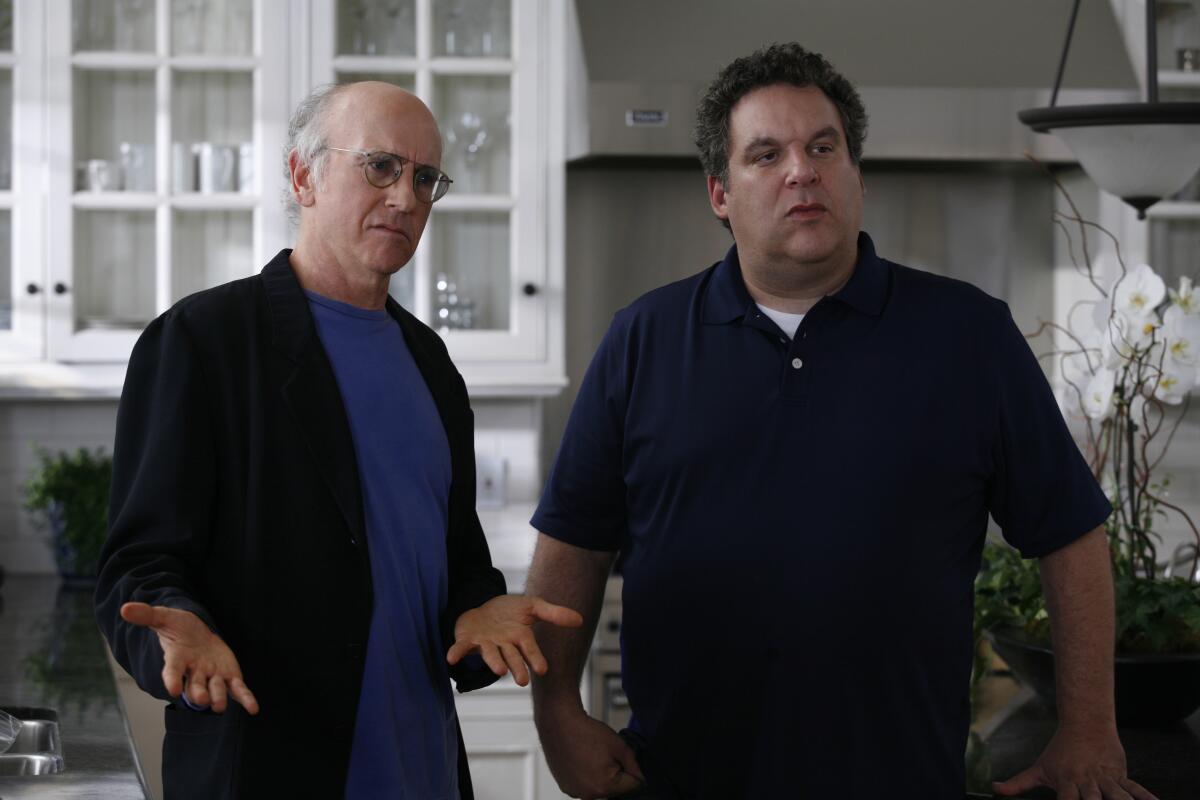 Two men in dark shirts stand in a kitchen looking confused.