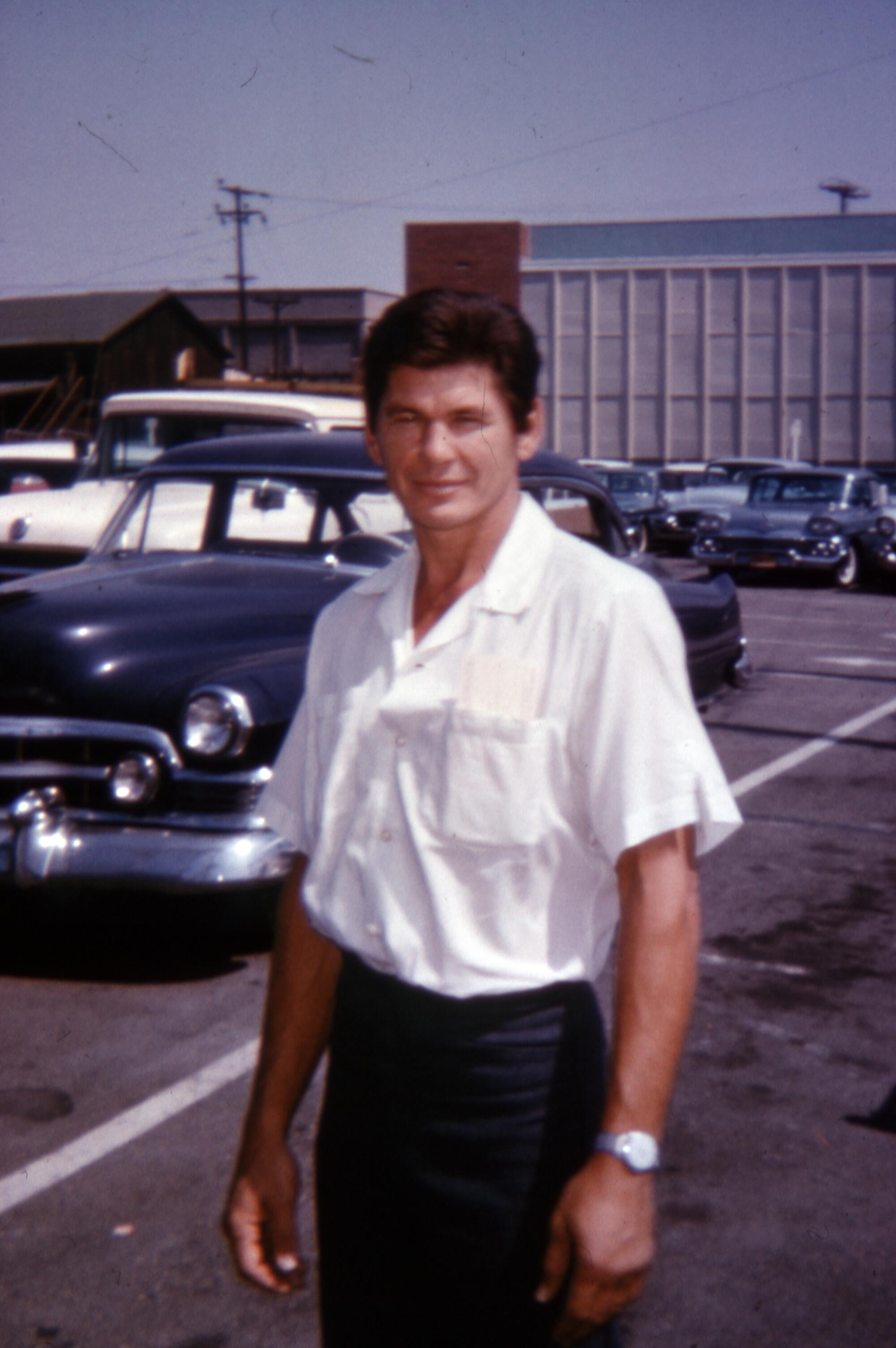 A dark-haired man in dark slacks and a white short-sleeve shirt stands in a parking lot filled with classic cars