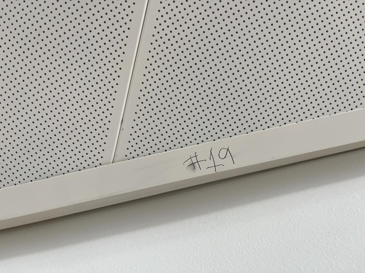 A close-up of of a perforated aluminum ceiling panel shows "#19" scrawled in pencil on the trim.