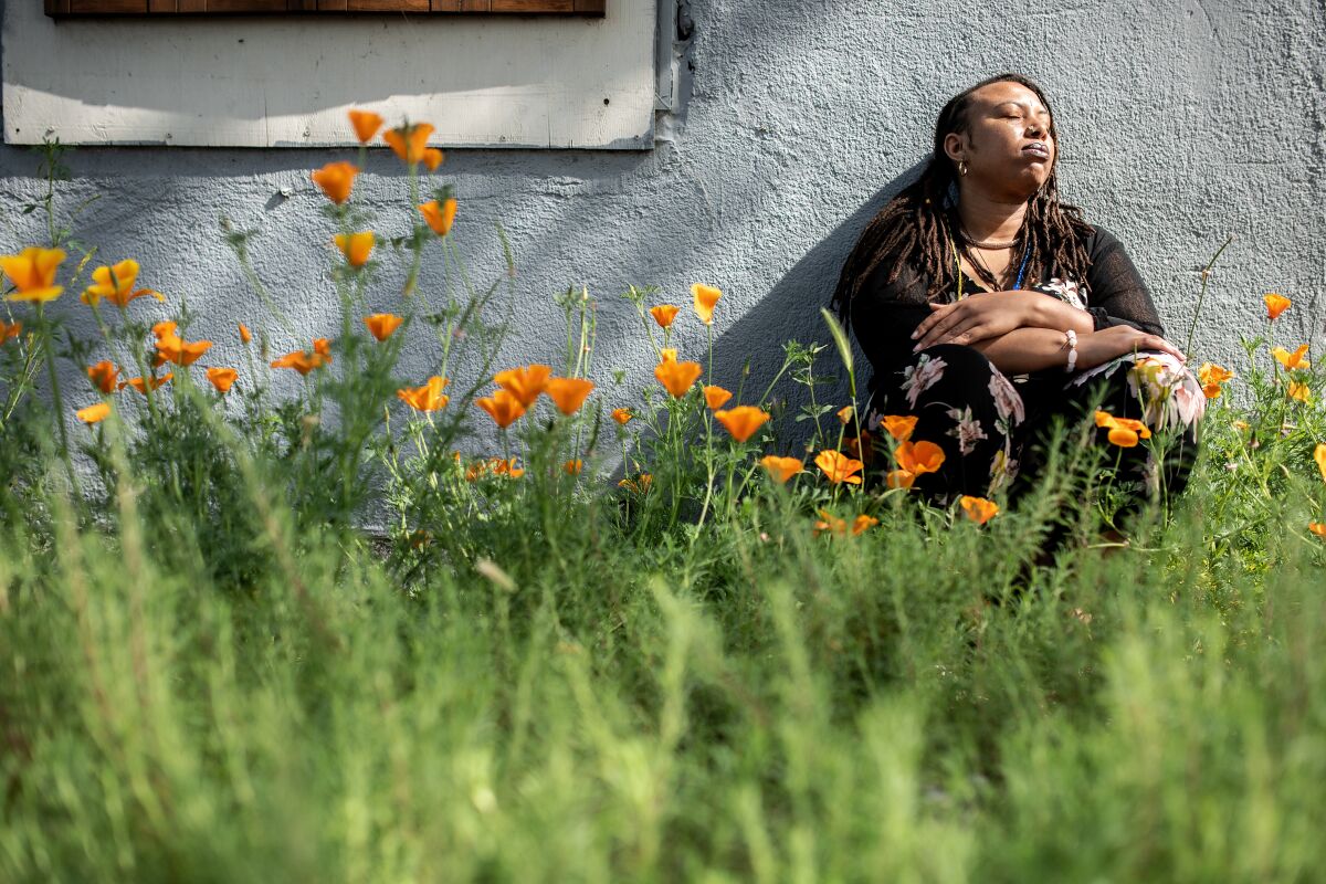 A person sits with her back against a wall at the Survivors Healing Garden, with orange flowers and grass around her.