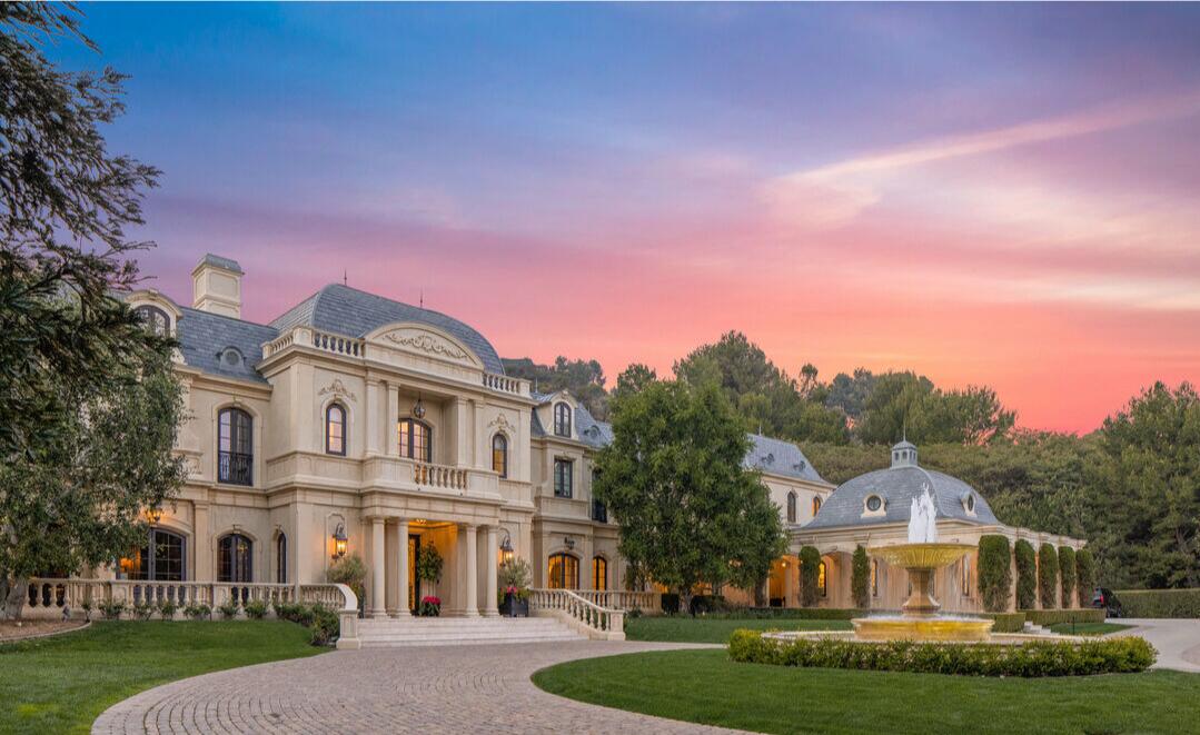 The exterior of the European-style mansion, driveway and lawn set against a pink and blue sky.