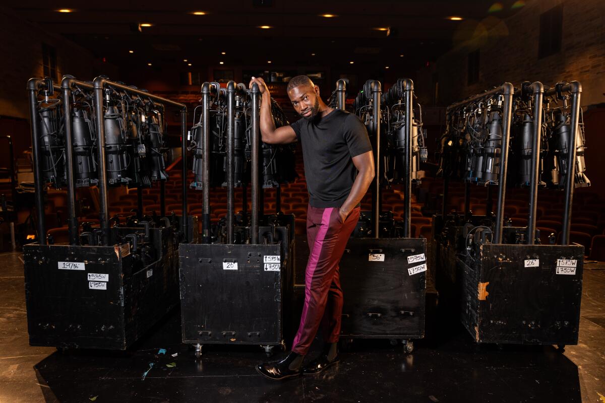 Tarell Alvin McCraney, in a black T-shirt and red pants with a pink stripe, stands in front of lights onstage.