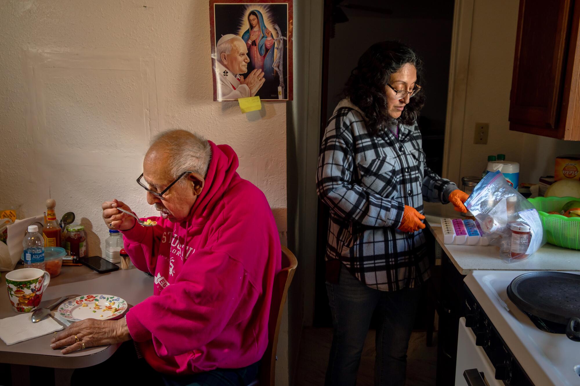 A woman stands at a kitchen counter refilling medication tray and a man sits at a kitchen table, eating.