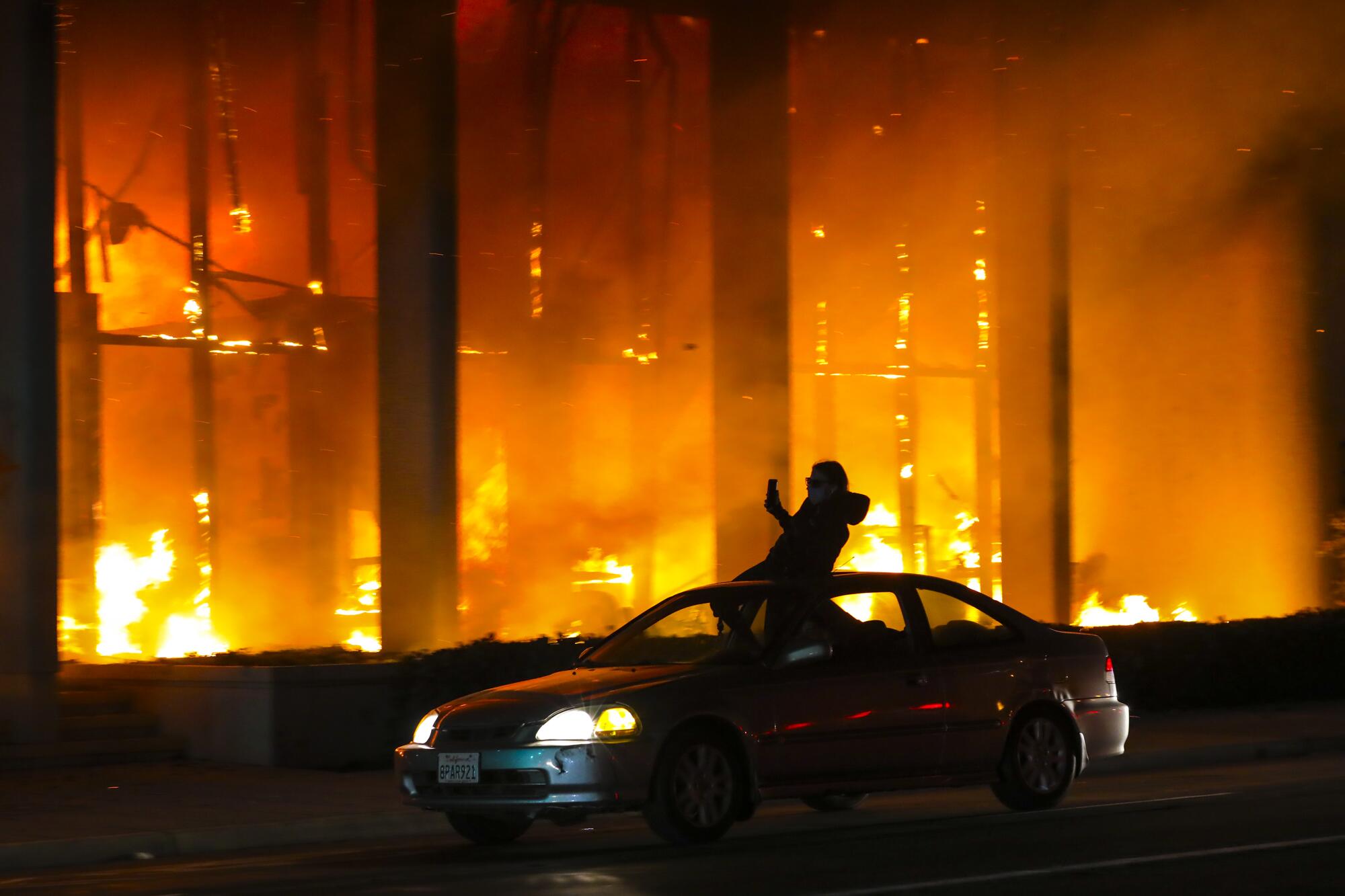 a car with someone standing out of the sunroof drives by a bank gutted by a raging fire