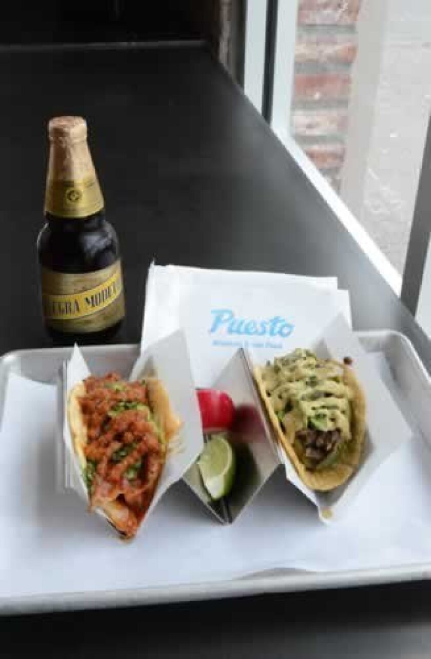 Two tacos served with a bottle of Negra Modelo beer. The taco on the left consists of chicken, cheese, pineapple, avocado, cilantro and tinga; the one on the right features beef, corn truffle, jalapeno, avocado, cilantro and onion.