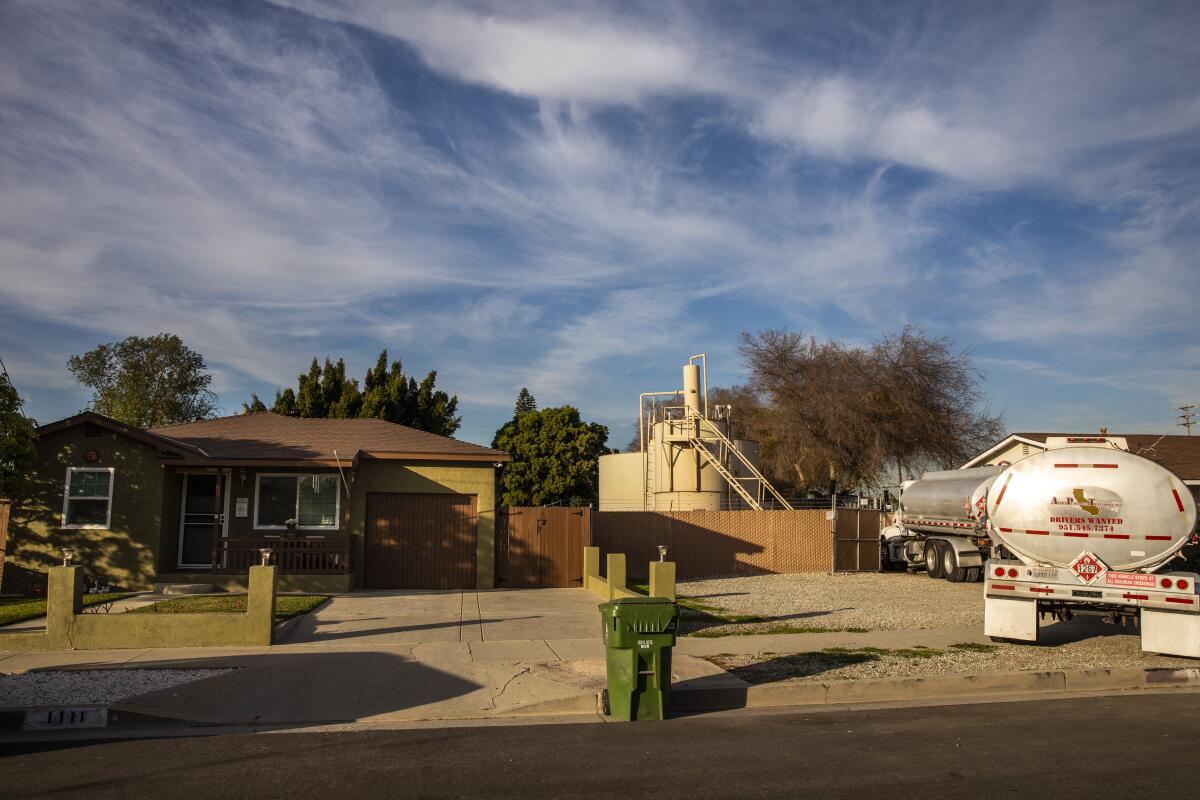 A green house sits to the left of oil infrastructure in a neighborhood as a tanker truck arrives