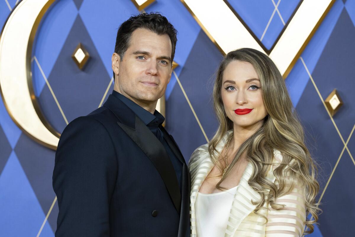 Henry Cavill, left, in a dark blue suit and Natalie Viscuso in a light-colored ensemble pose in front of a dark backdrop