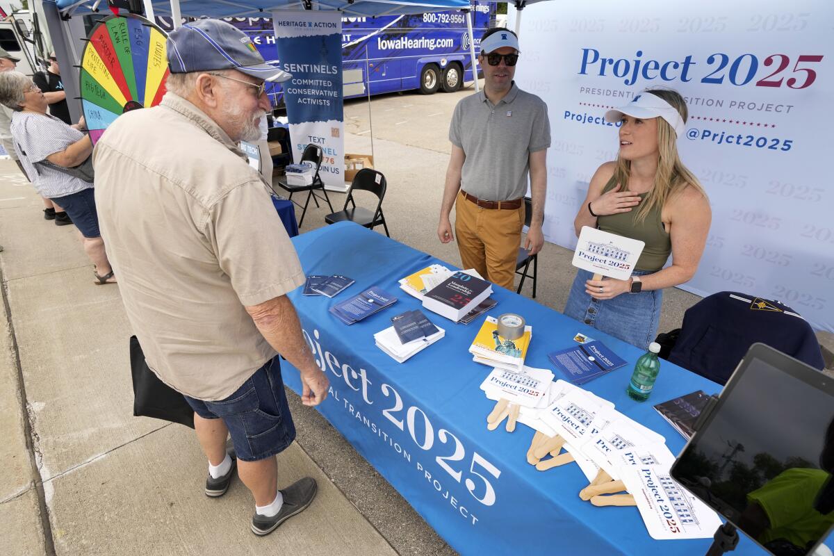 A man in a cap, khaki shirt and dark shorts speaks to people behind a table bearing the words Project 2025