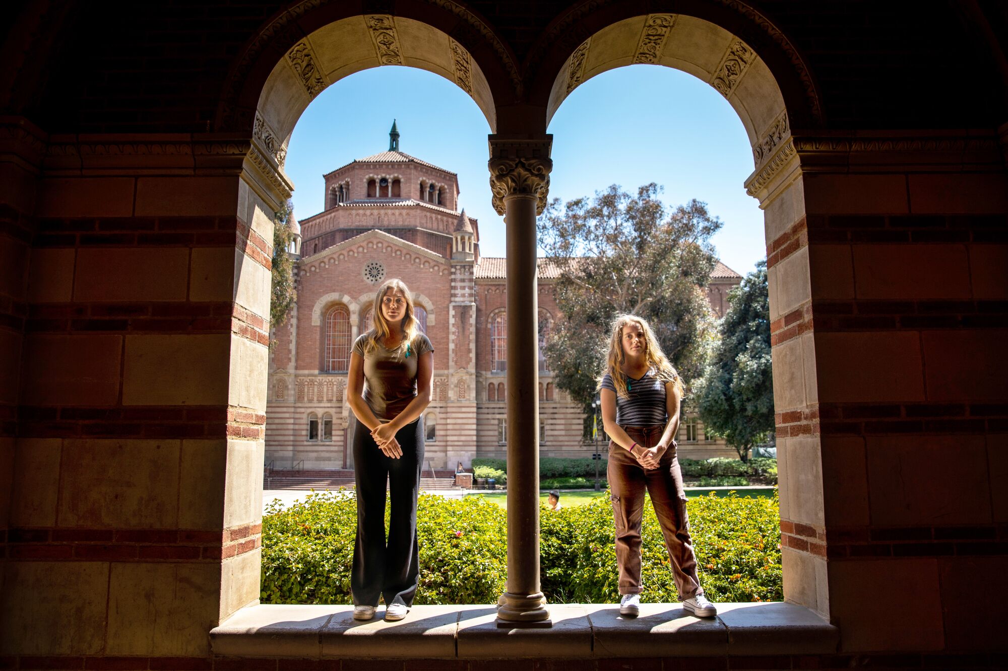 Two young women stand on a ledge of an archway at a university campus, looking firmly at the camera.