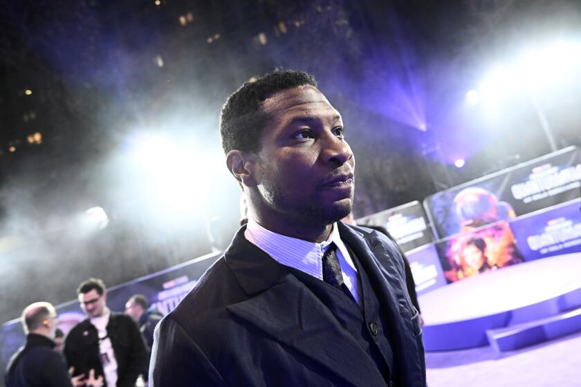 Jonathan Majors looking ahead, walking in black suit with white shirt and black tie at movie event