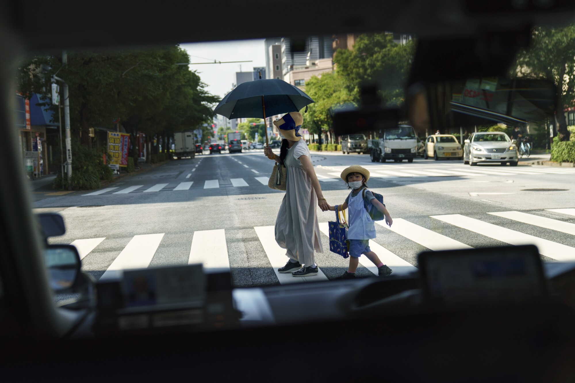 A woman with an umbrella holds a child's hand on a sunny street.