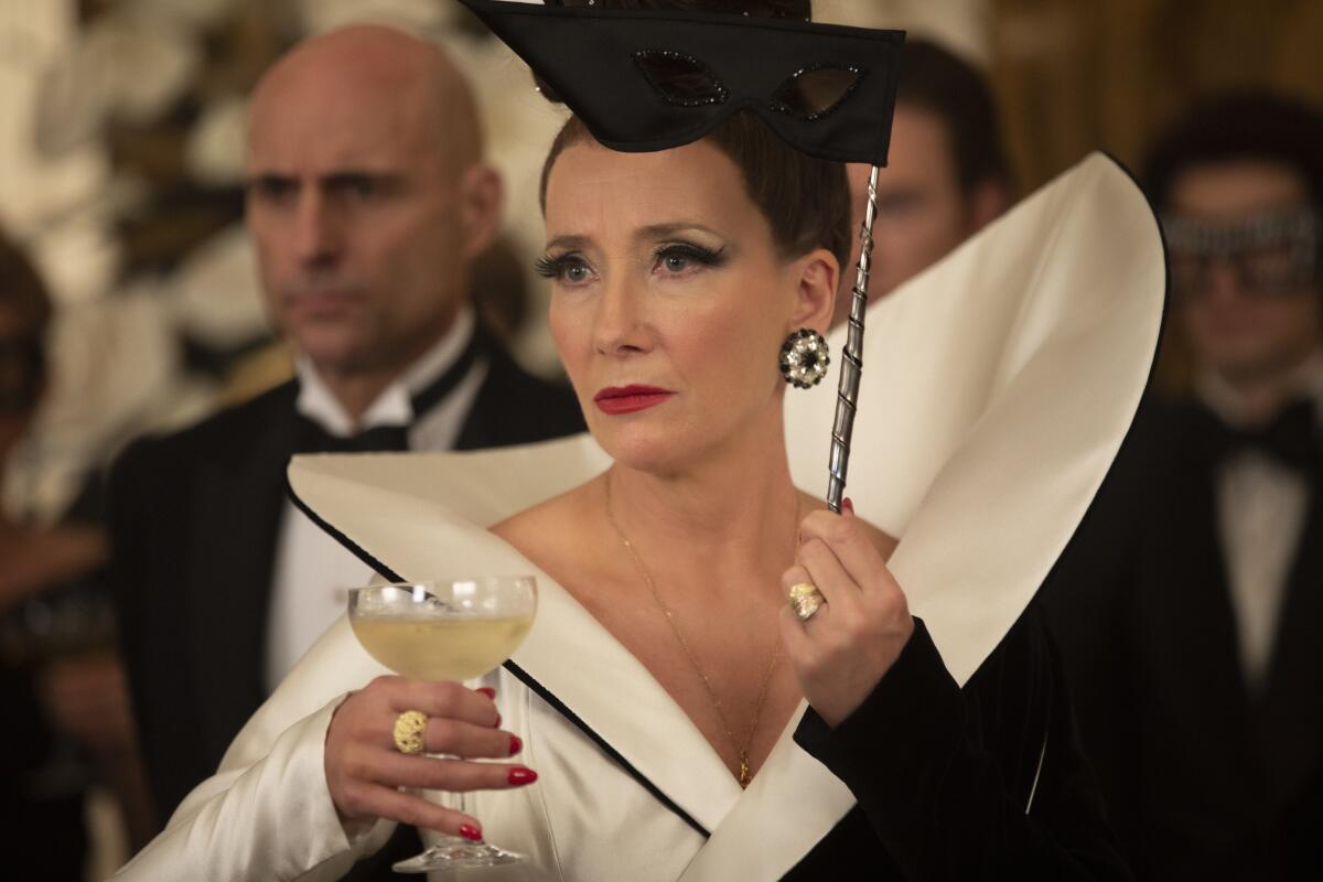 Emma Thompson as the Baroness holds a wine glass and a mask with a man in a tuxedo behind her.