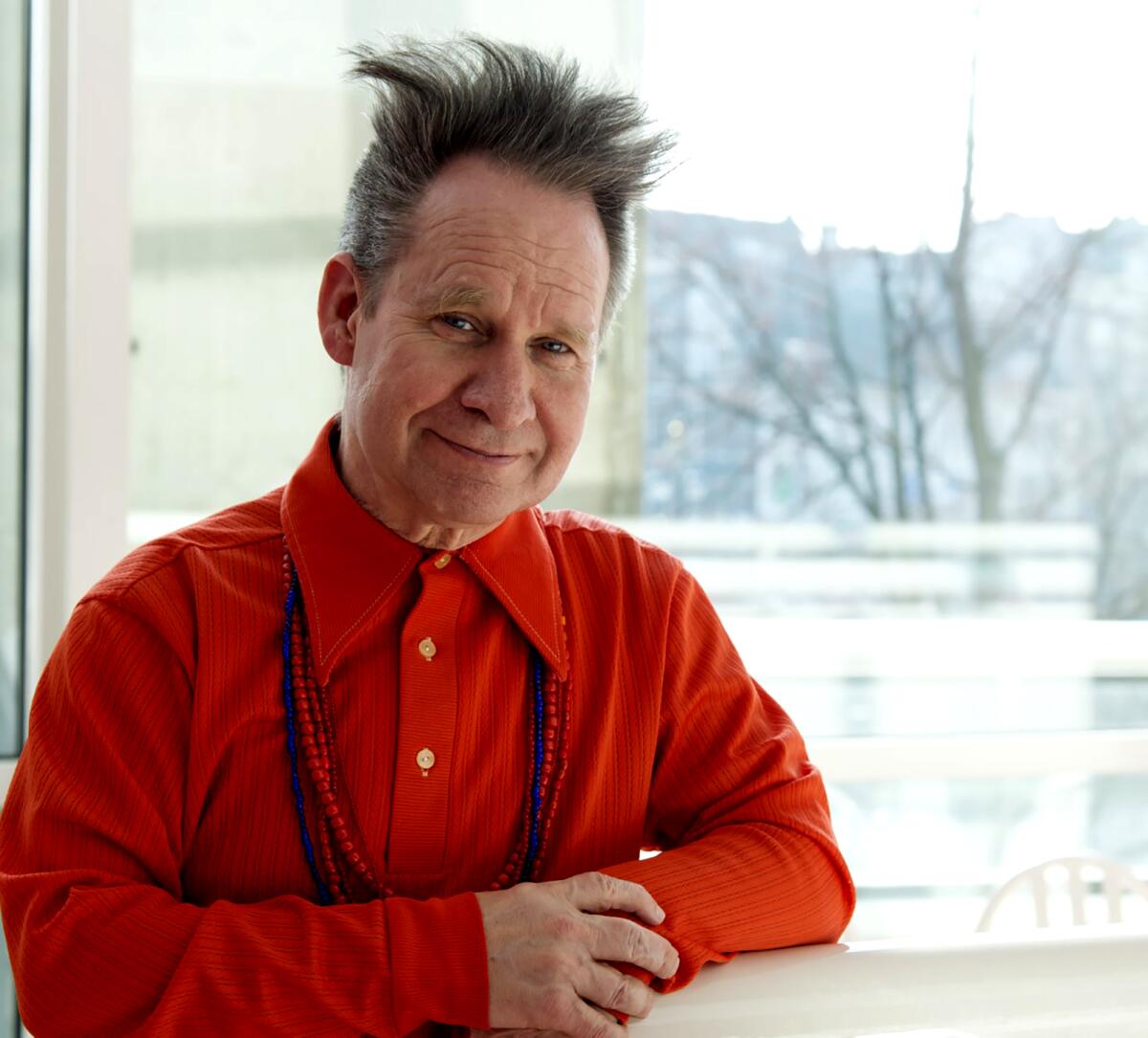 A smiling man in a red button-front shirt with beaded necklaces sits at a table in front of a sunny window.