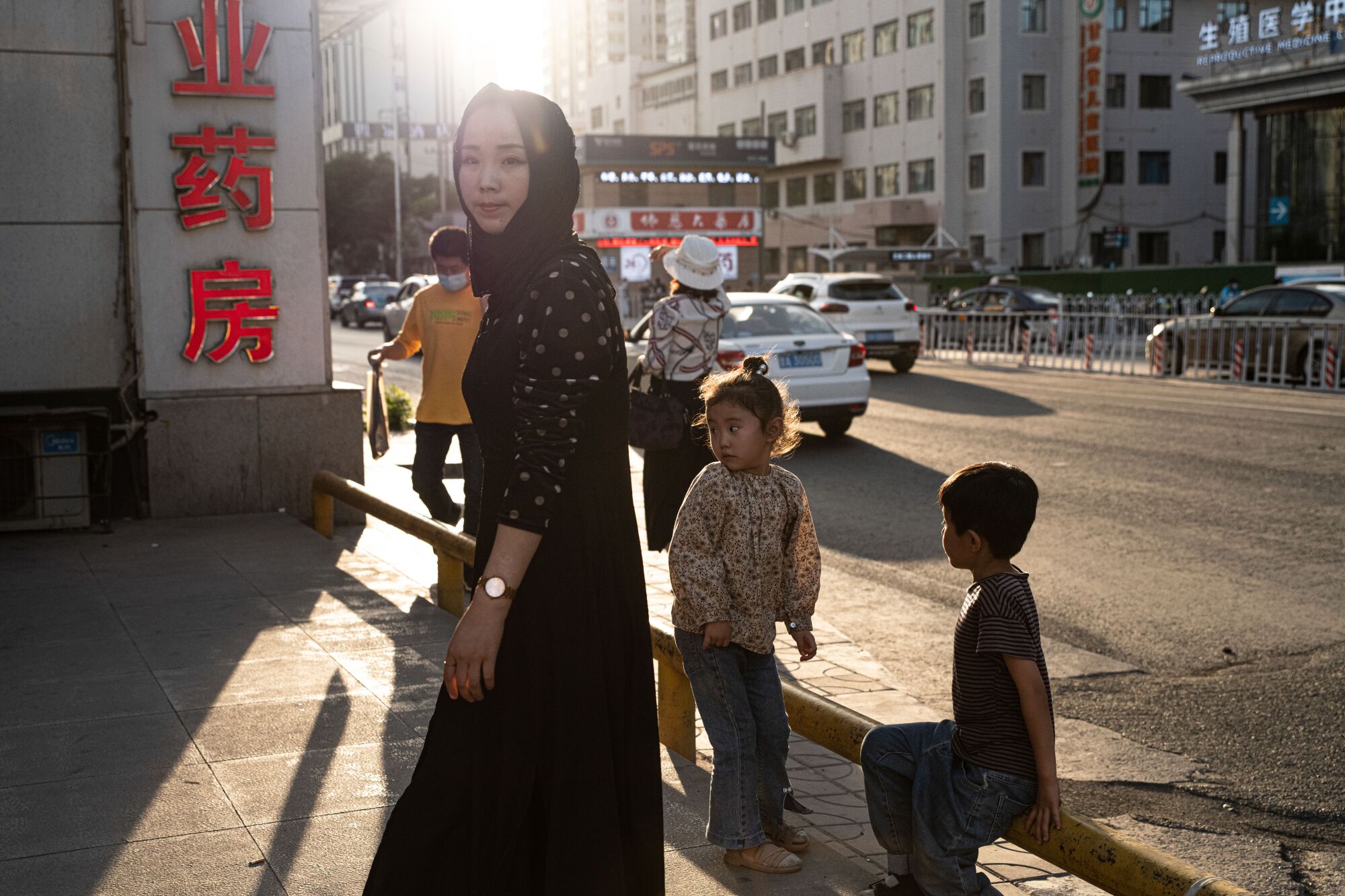 A woman in a dark head scarf and dark clothing stands near two children and a building with red Chinese characters