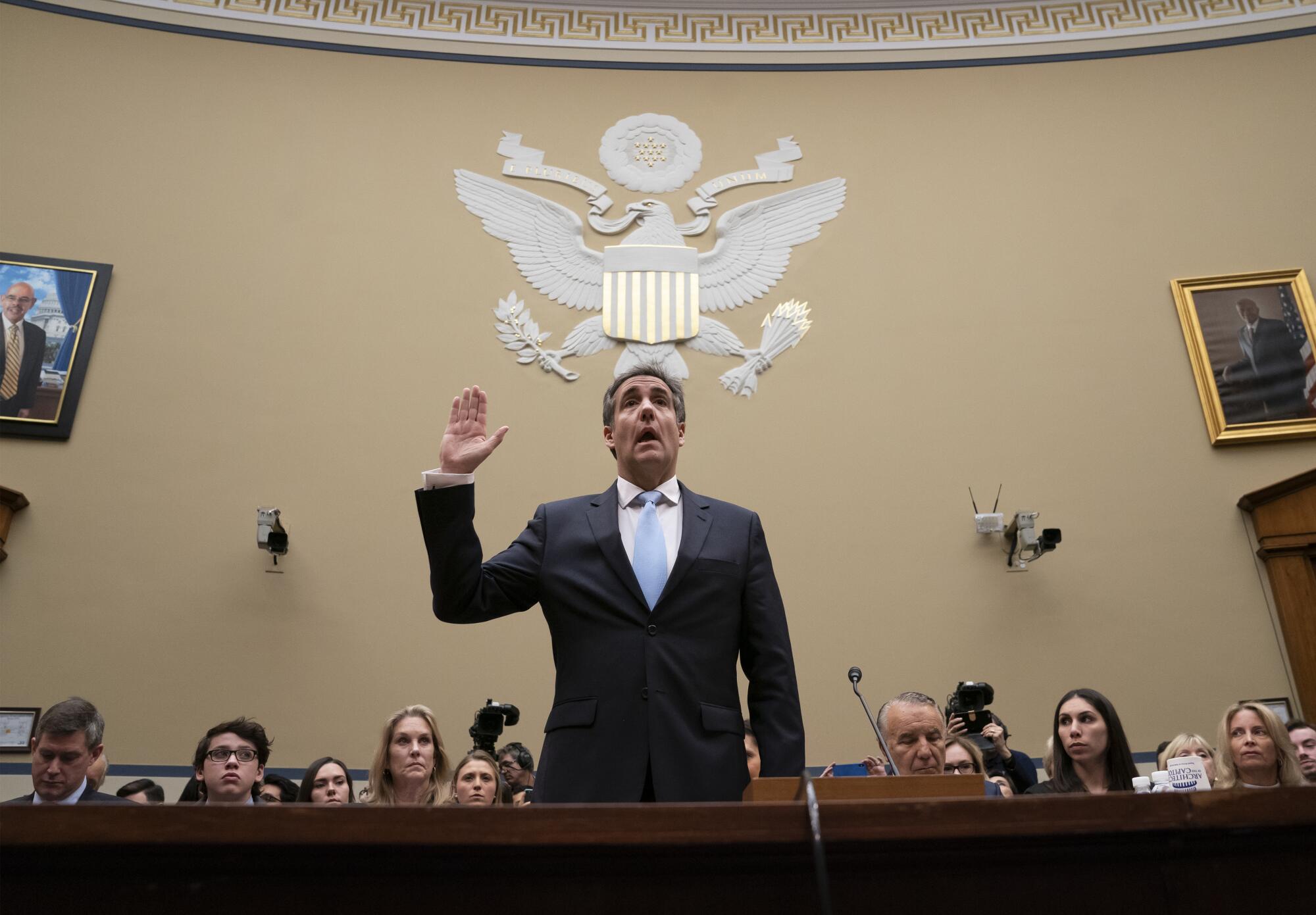 گروه نشسته اند و مایکل کوهن در حالی که دست راستش را برای ادای سوگند بالا آورده است، زیر تصویر عقاب روی مهر مجلس نمایندگان ایستاده است.