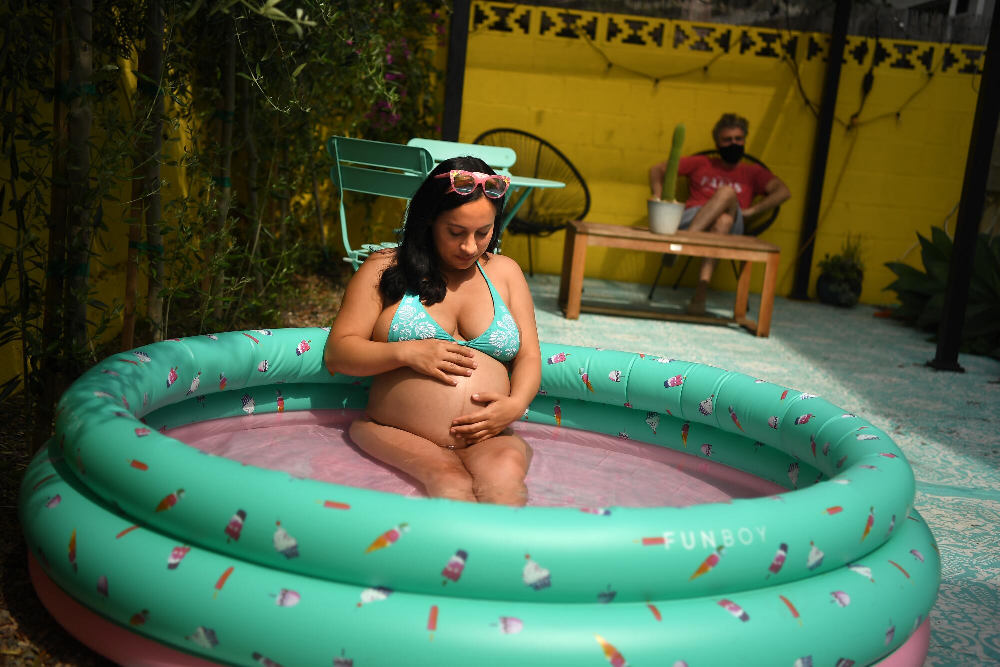 A pregnant woman in a green bikini has her hands on her belly while sitting in an inflatable pool