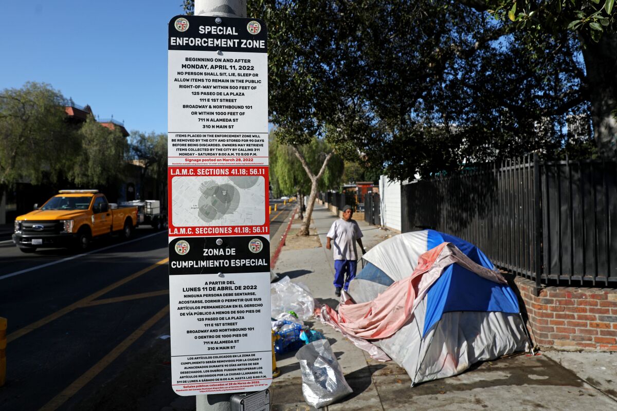 A homeless man is shown in the background near a sign for a new ordinance in Los Angeles.
