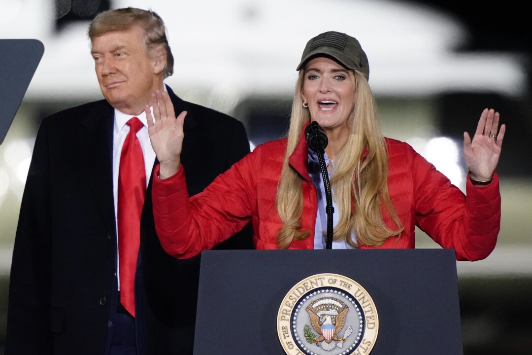 President Trump smiles as Kelly Loeffler speaks at a podium.