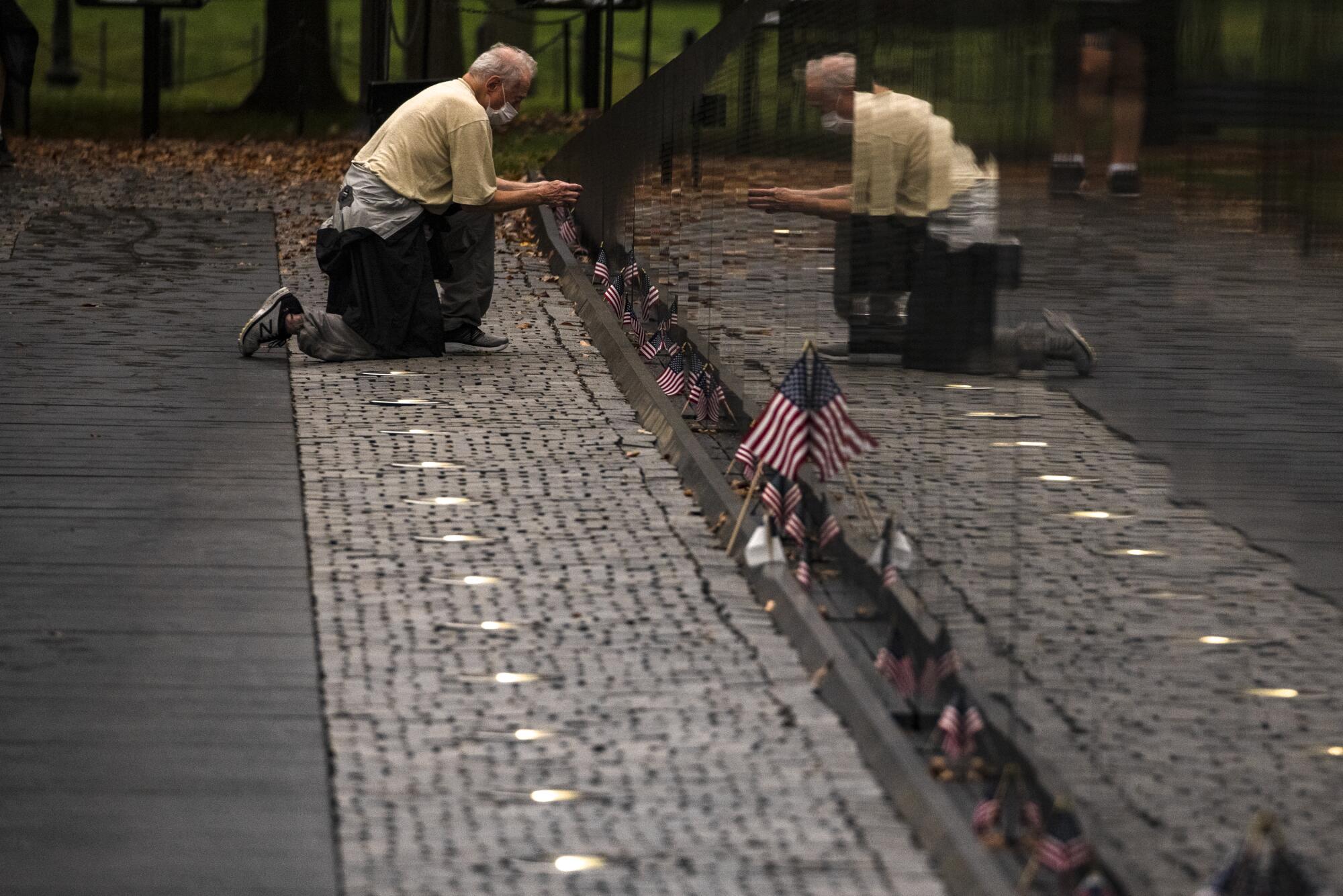 A man in a face mask kneels and peers at the Vietnam Veterans Memorial Wall.
