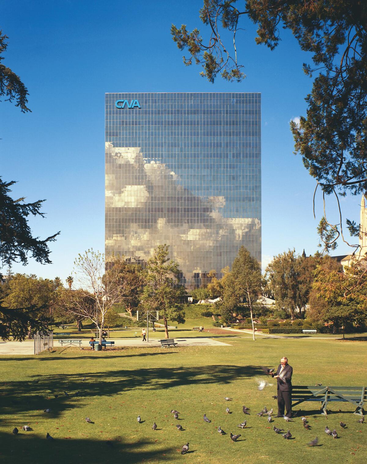 Clouds and sky are reflected on a glassy tower that rises above a park where a man feeds pigeons