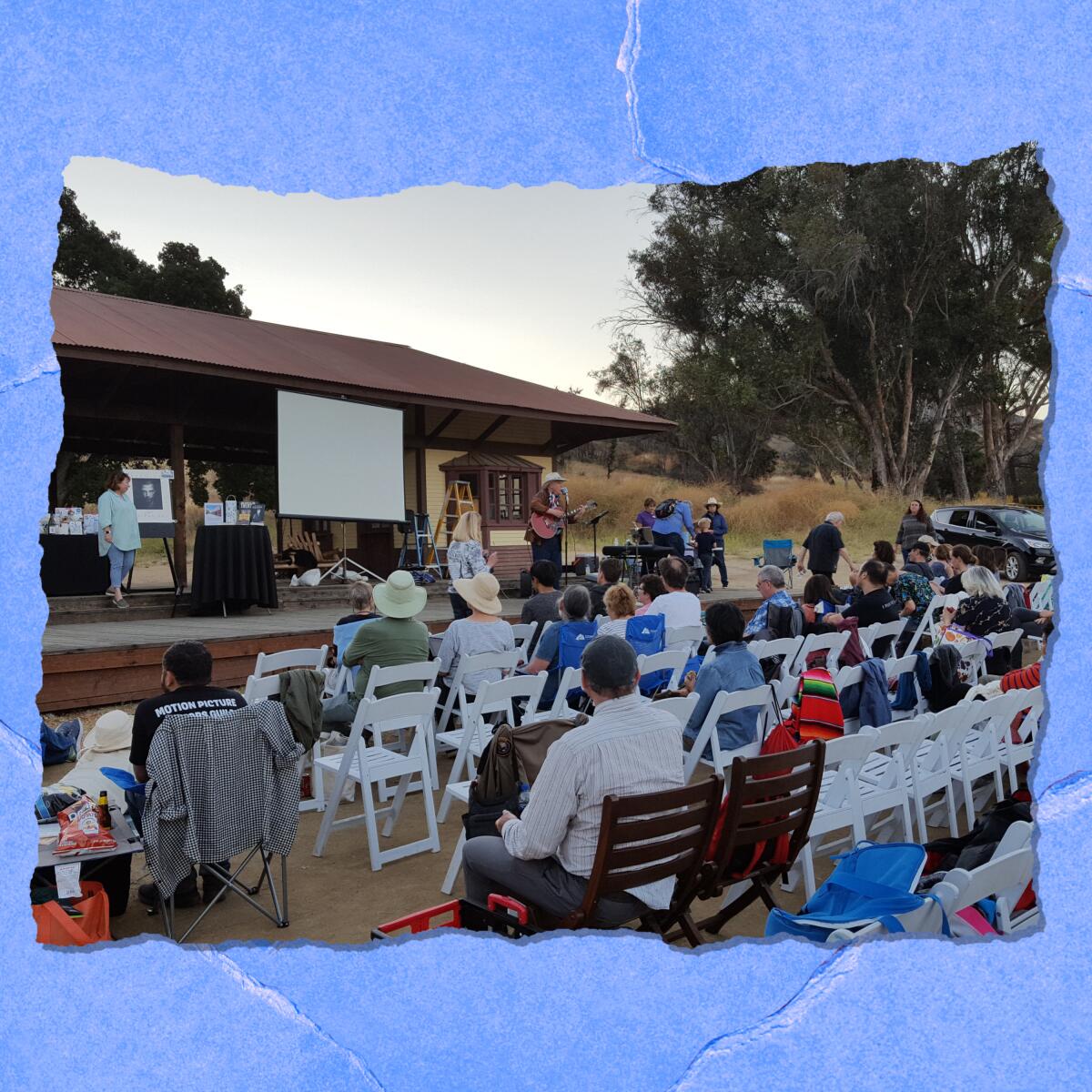 People sit on folding chairs outdoors in front of a screen.