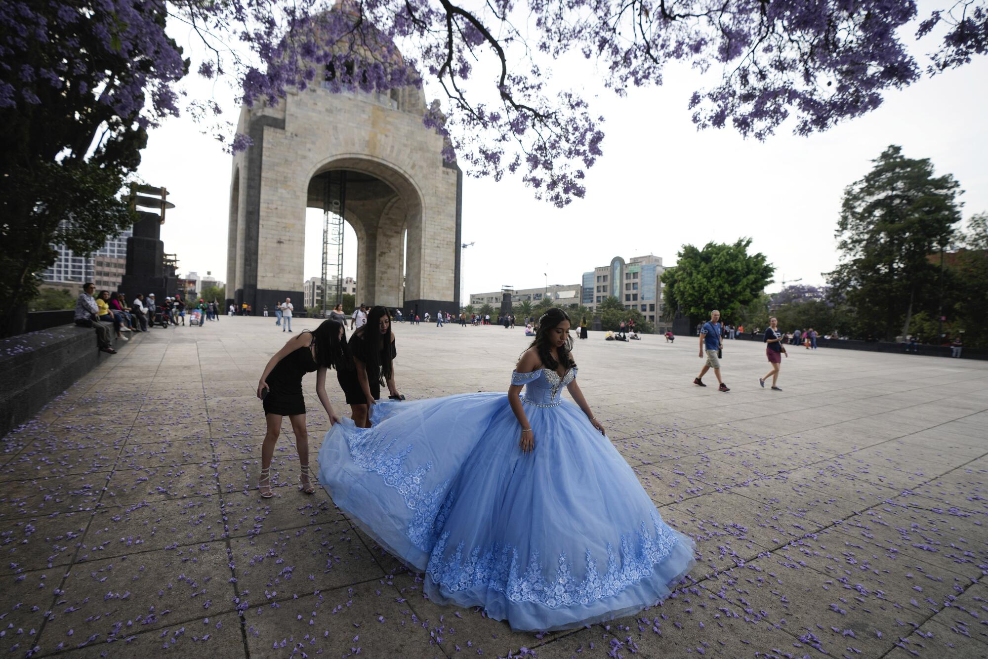 两个人穿着女孩的蓝色礼服调整火车，背景是一座巨大的纪念碑，头顶是紫色的花朵。