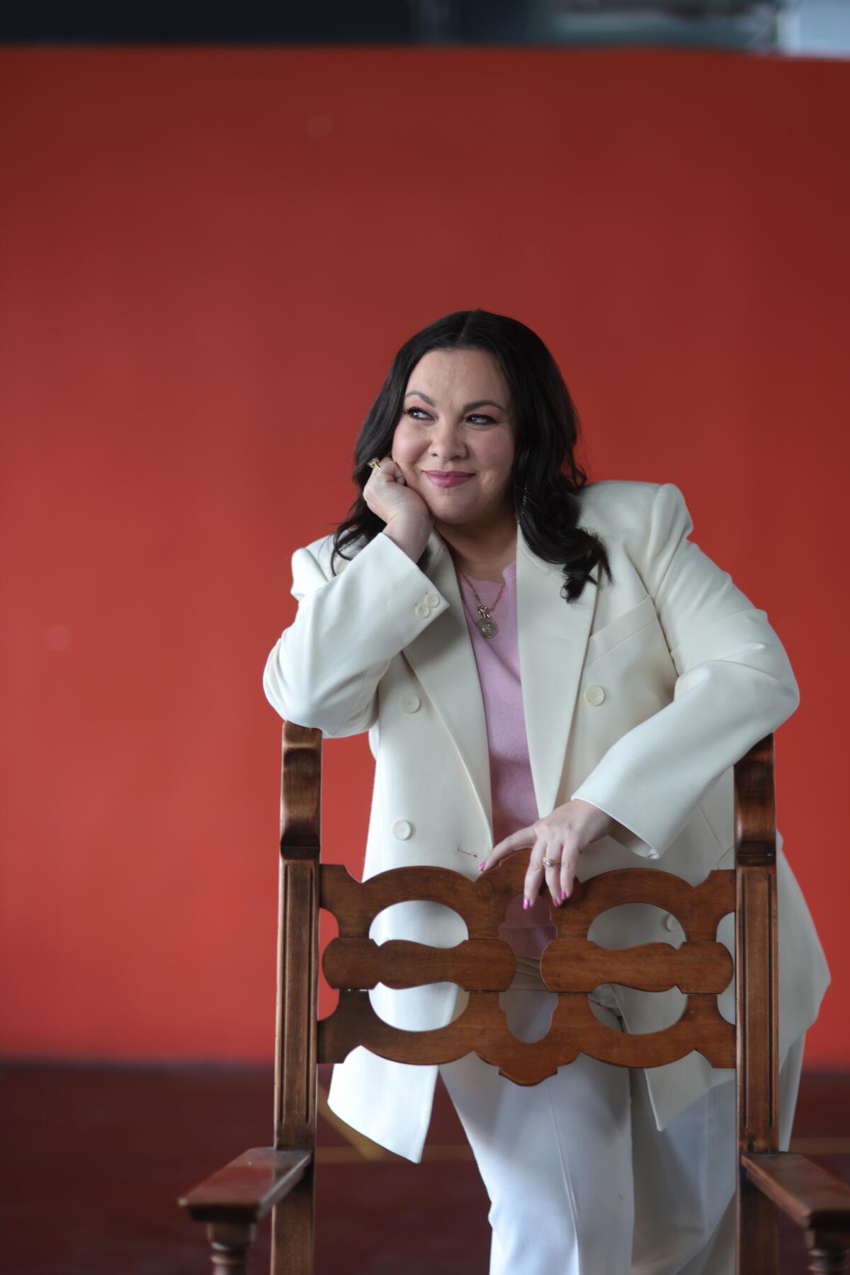 A smiling woman in a white suit leans against the back of a chair