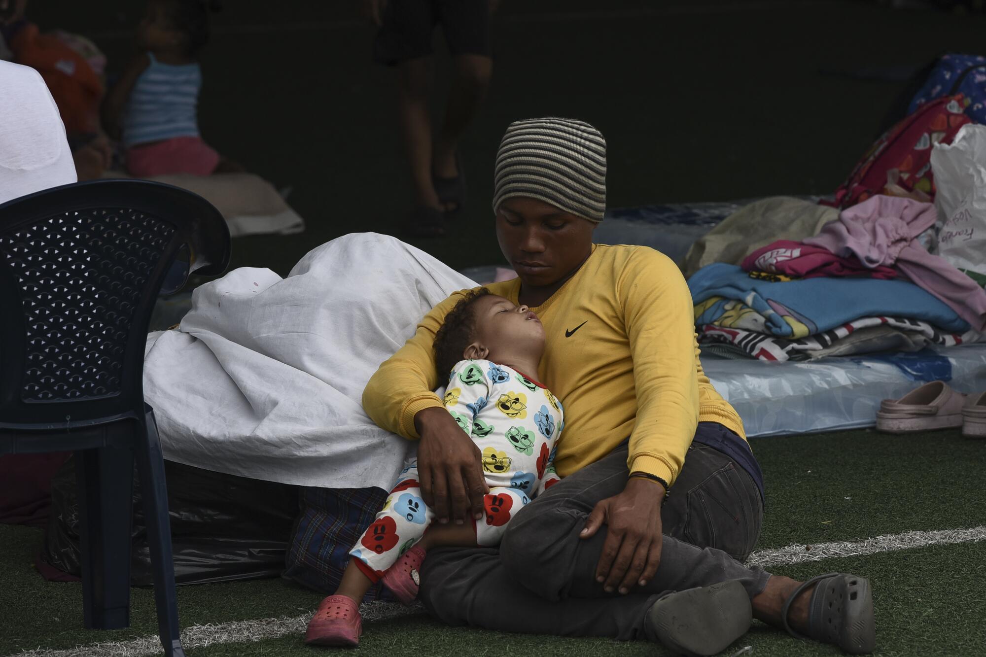 A person in a knit cap, yellow shirt and dark pants holds a sleeping toddler while lying on the ground