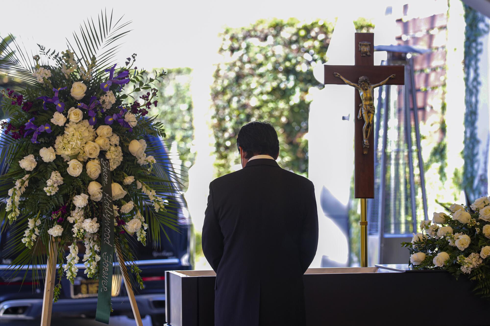 A man, seen from behind, stands at an open casket flanked by floral arrangements and a crucifix